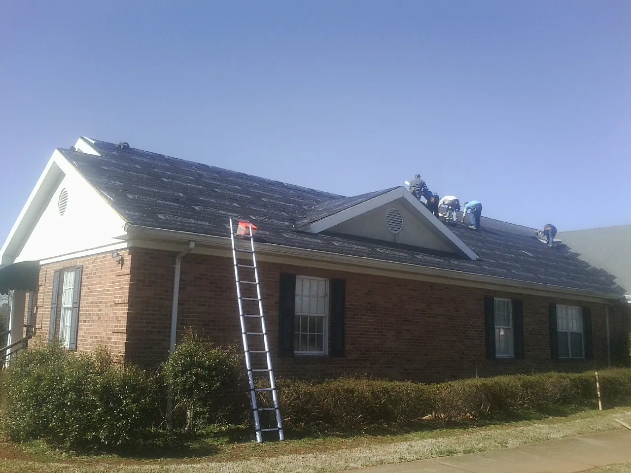 Crew tearing shingles off a brick ranch roof with a ladder set — Emergency Roof Repair work by our crew in Mc Arthur