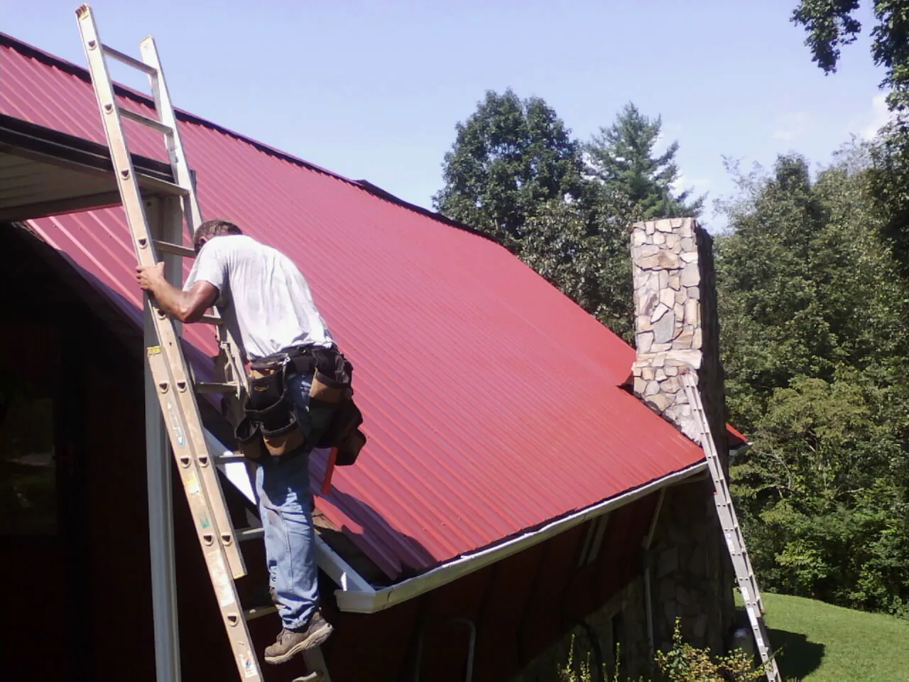 Roofer on a ladder beside a red metal roof and stone chimney — Soffit & Fascia Repair by Roofing Brothers in Mc Arthur